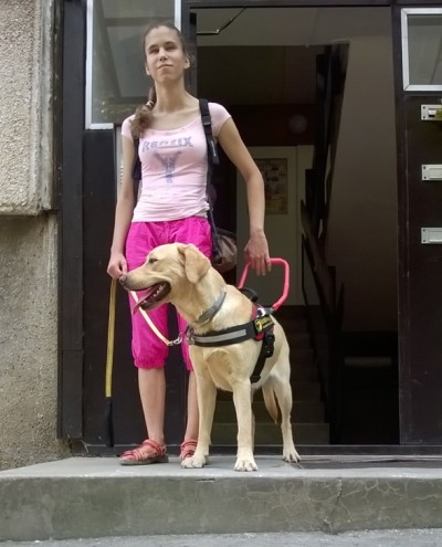 A képen egy fiatal lány áll a lécső tetején vakvezető kutyájával, aki egy sárga labrador.