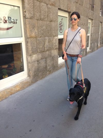 A képen egy látássérült hölgy fekete vakvezető kutyájával megy a járdán.