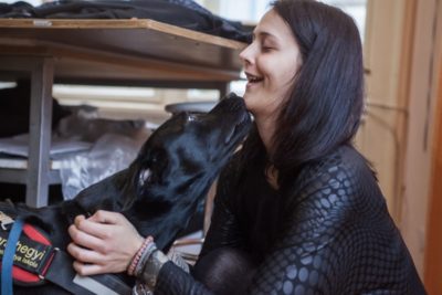 A képen Frutti a vakvezető kutya, aki egy fekete labrador és puszit ad gazdájának, Zsuzsinak.