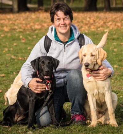 A képen Piroska egy parkban féltérdre ereszkedve guggol, miközben jobb kezével egy fekete, bal kezével egy sárga labrador kutyust ölel