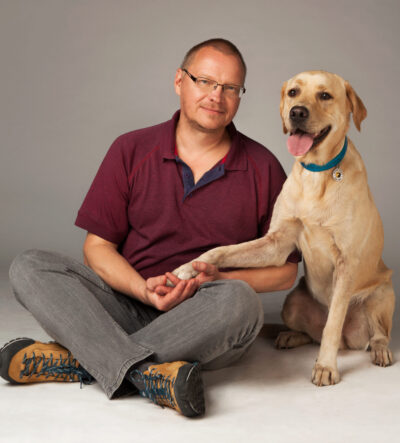 Tamás és Trafi A képen Tamás egy stúdióban törökülésben ül a földön. A kezeit az ölében tartja, amire egy sárga labrador (Trafi) mellette ülve ráteszi a jobb mellső mancsát