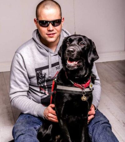 A képen Zoli a földön ül kinyújtott lábakkal, a fekete labrador kutya a két lába között mosolyogva ül